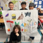 Three kids holding up a poster with one sitting in front of it